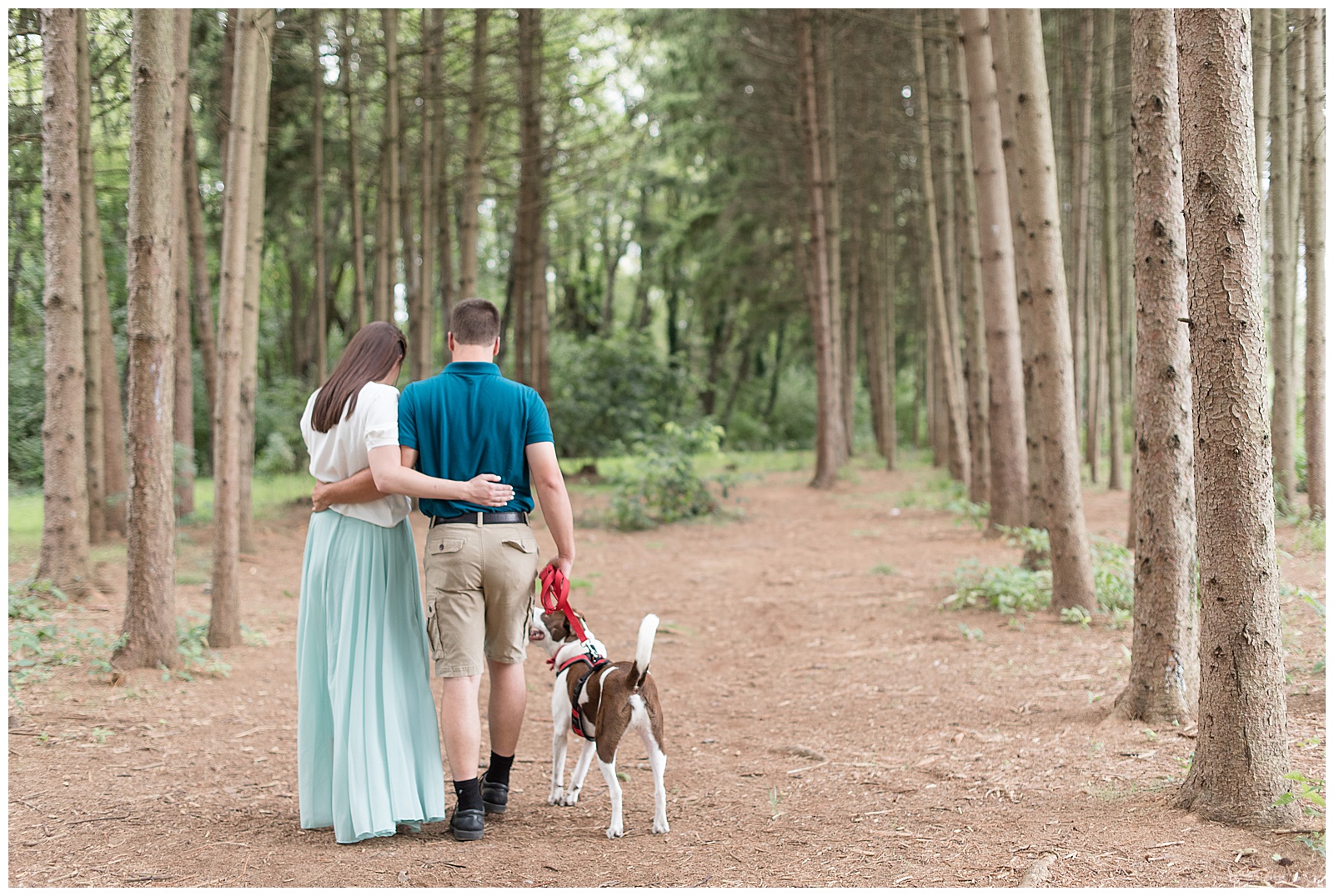 the couple is on the left side of the photo hugging with arms wrapped behind each other with their backs to the camera and the girl is on the left and the guy is on the right and they are looking down at their dog which is to the right of the guy as he holds the dog's leash in his right hand on the path with the rows of evergreen trees surrounding them at Overlook Park in Lancaster, PA