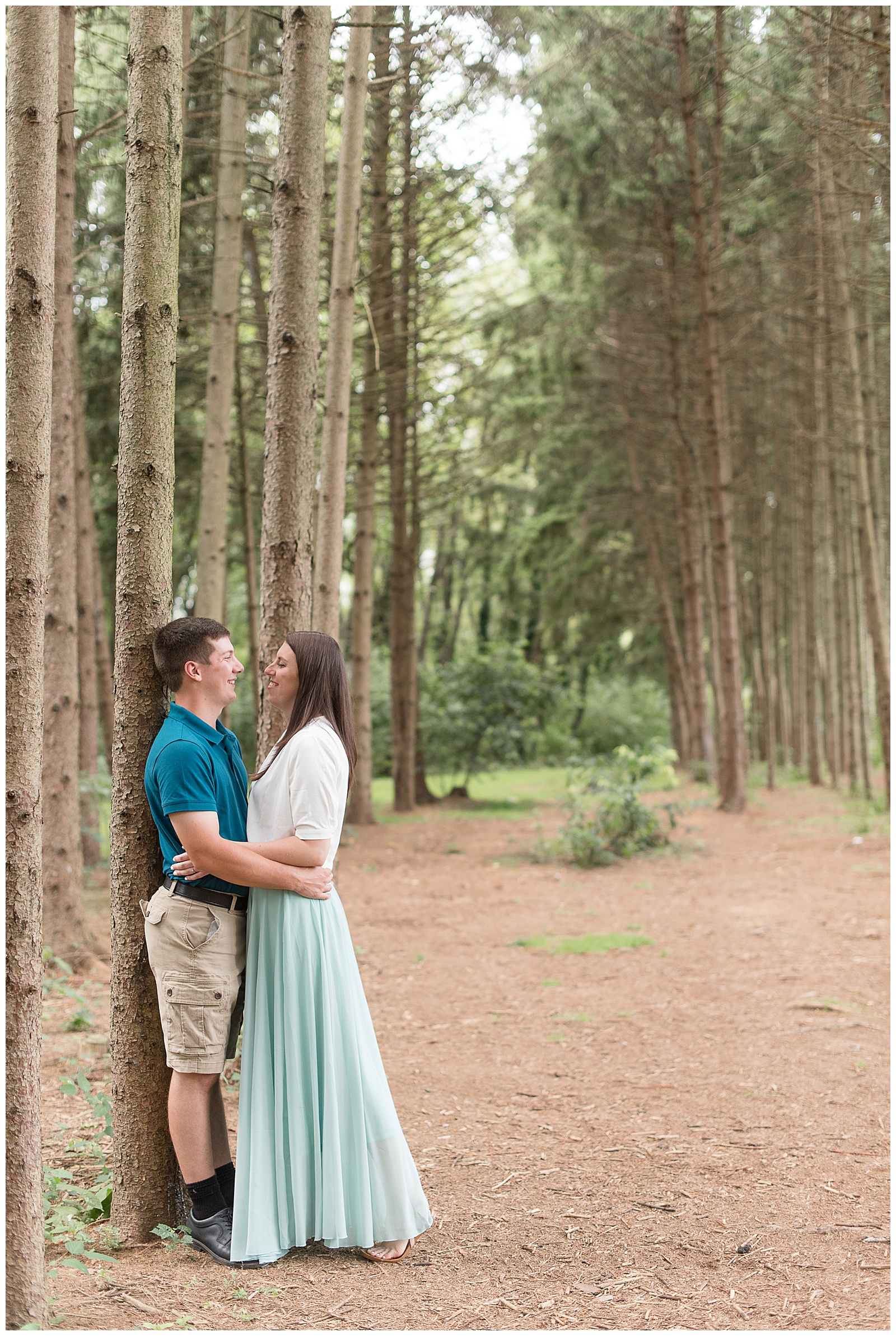 the couple is standing close together face-to-face looking at each other and smiling as the guy is on the left leaning against one of the evergreen trees and the girl is on the right and their arms are around each other's waist while standing on the path surrounded by the rows of evergreen trees at Overlook Park in Lancaster, PA