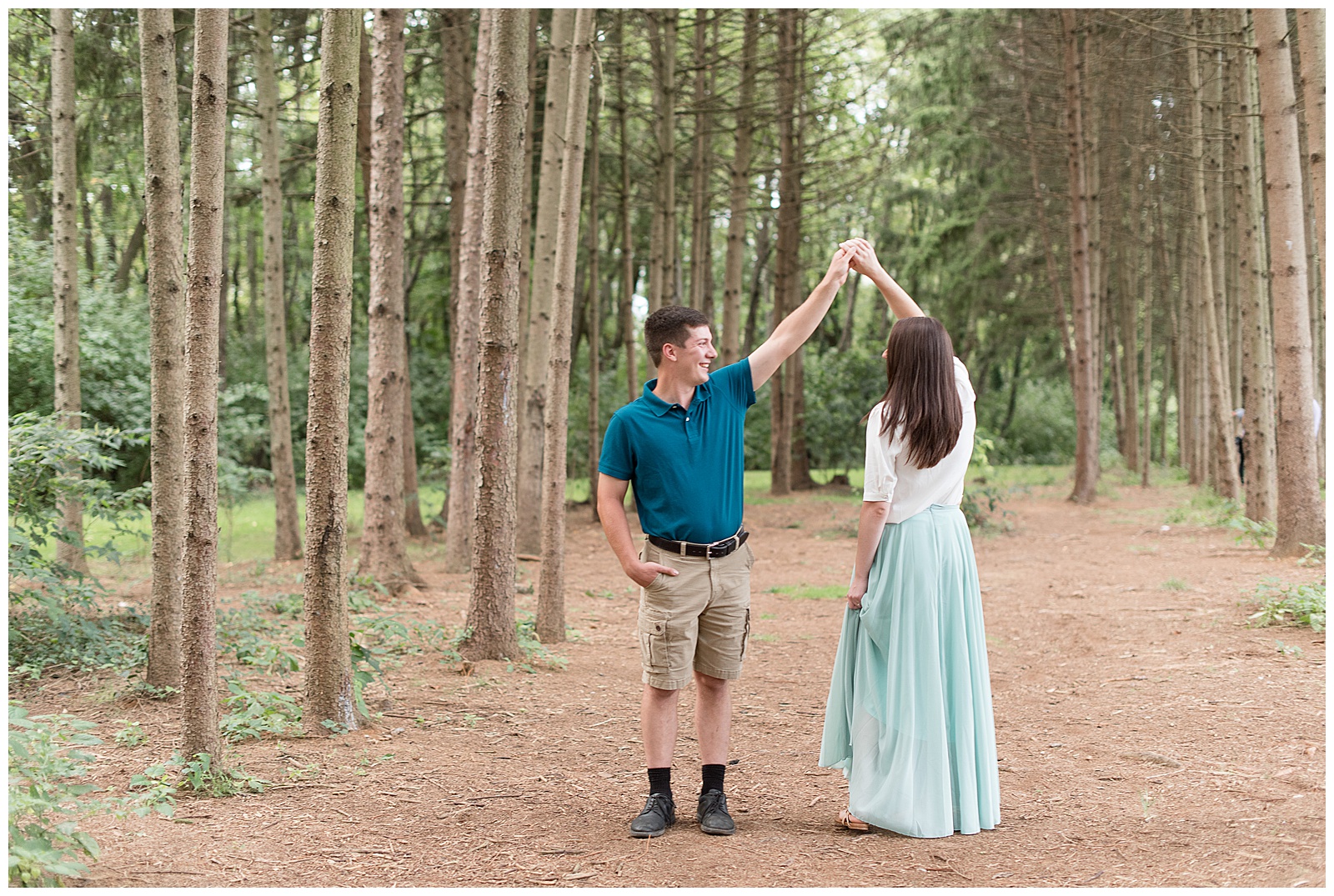 the couple is standing on a path in between rows of evergreen trees shading them and the guy is on the left with his right hand in his pocket and his left arm held high twirling the girl underneath their raised arms and he is looking towards the girl smiling and the girl's back is to the camera as her left hand is lightly grasping her mint green skirt at Overlook Park in Lancaster, PA
