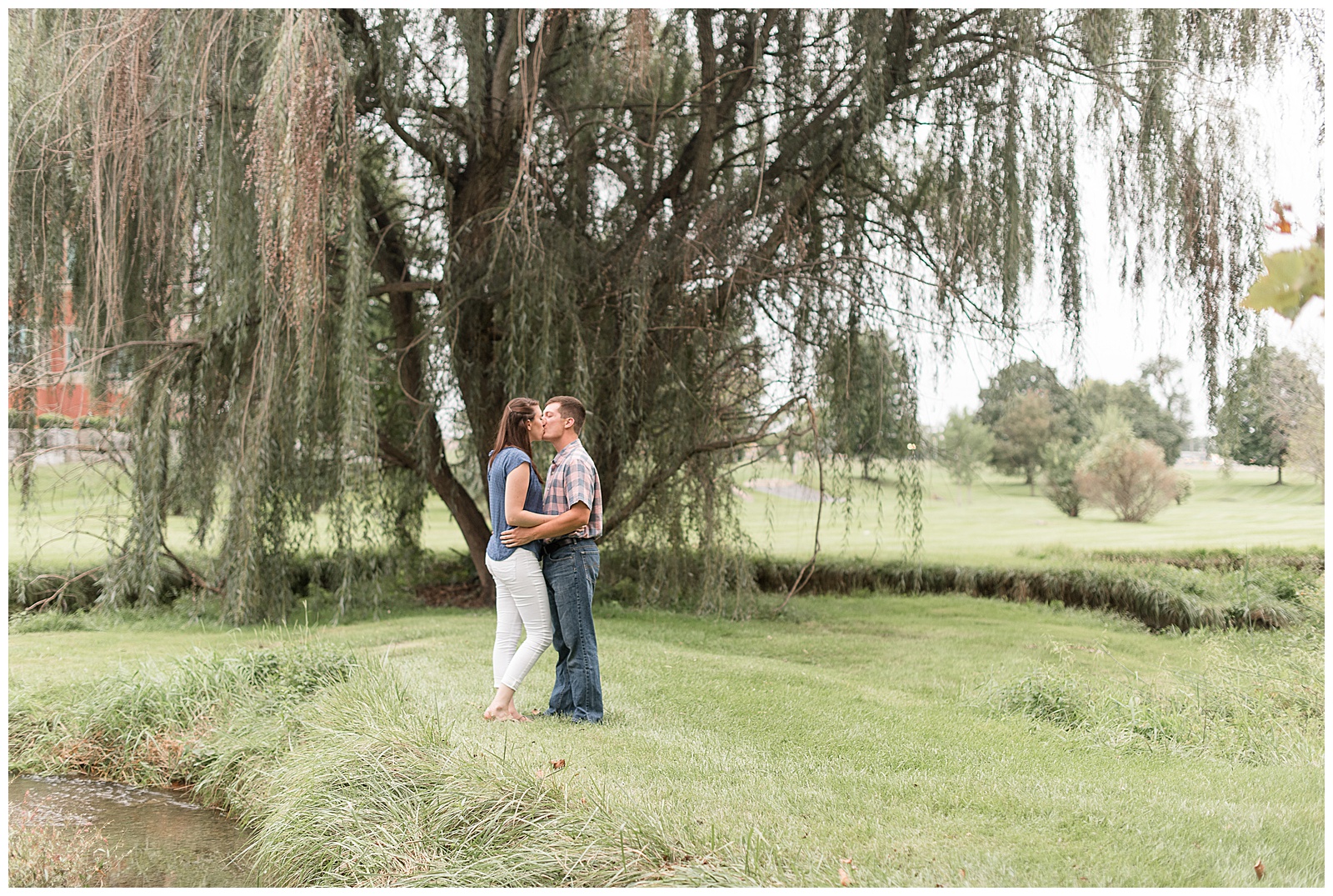 couple is standing in grassy field with large willow tree behind them on an overcast evening with the girl on the left and the guy on the right with their arms around each other and they are kissing at Overlook Park in Lancaster, PA