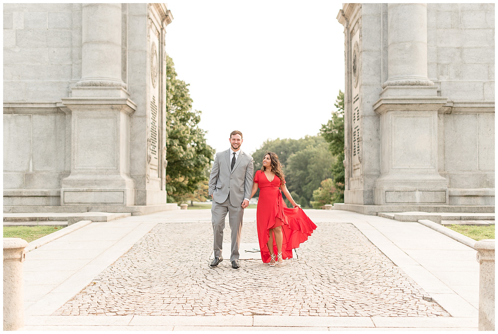 couple is under large archway with trees behind them and they are walking towards the camera with the guy on the left and his right hand in his pocket and the girl is on the right with her right arm wrapped around his left arm and her left arm is holding her red dress out to the side and they are looking at the camera and smiling at the National Memorial Arch in Valley Forge, PA