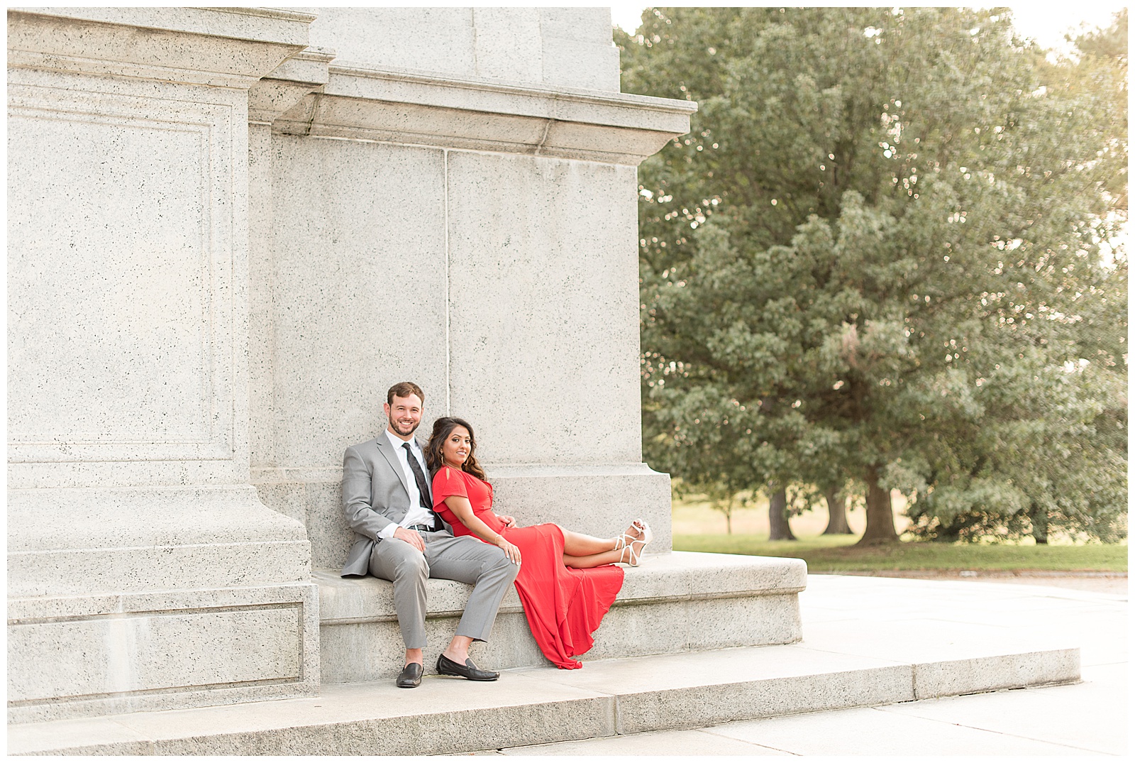 couple is sitting on archway bench with the guy on the left and the girl on the right and he is resting his right hand on his knee with his left arm around the girl and his feet are on the ground while the girl is on the right sitting with her legs extended out along the bench and feet are crossed as her red dress drapes over the bench and her right hand rests on his left knee and they are looking at the camera smiling with trees behind them to the right at the National Memorial Arch in Valley Forge, PA