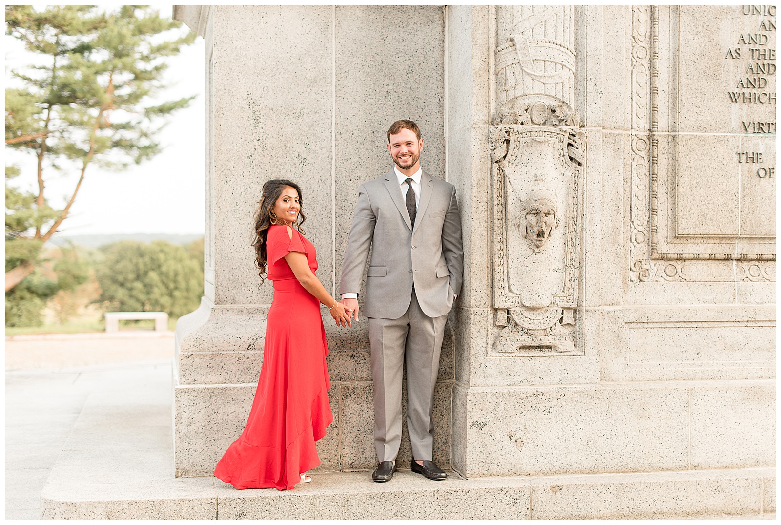 couple is standing in corner of large archway with the girl on the left and guy on the right and her body is turned towards the guy with her right hand holding his left hand as she looks directly at the camera smiling and the guy has his left hand in his pocket as he looks at the camera smiling at the National Memorial Arch in Valley Forge, PA