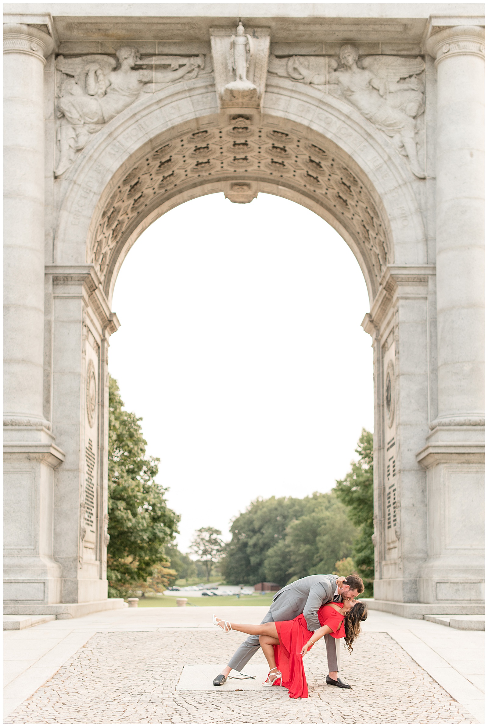 the couple is standing on the walkway under the archway and the guy is dipping the girl backwards with her left foot extended outward and he is slightly behind her on the left and there are trees in the distance behind them at the National Memorial Arch in Valley Forge, PA