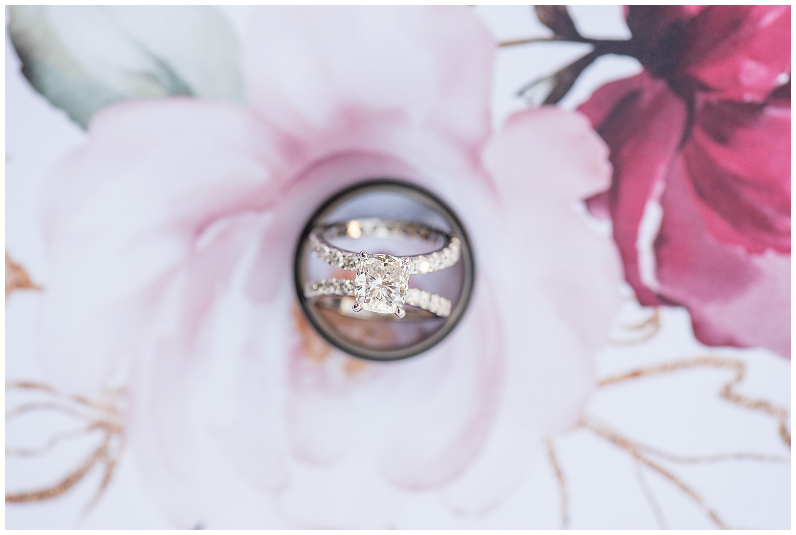 close up photo of brides wedding and engagement rings resting inside the groom's wedding band on light pink and maroon floral background at Riverdale Manor in Lancaster, PA