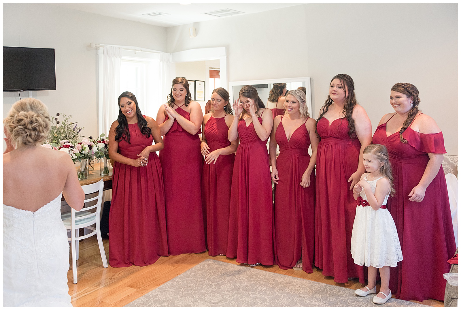 bride stands in lower left corner of photo with her back to the camera in bridal sweet with her row of bridesmaids turned towards her smiling and wiping tears as they see her in her gown and the flower girl standing in front the the bridesmaid on the far right at Riverdale Manor in Lancaster, PA