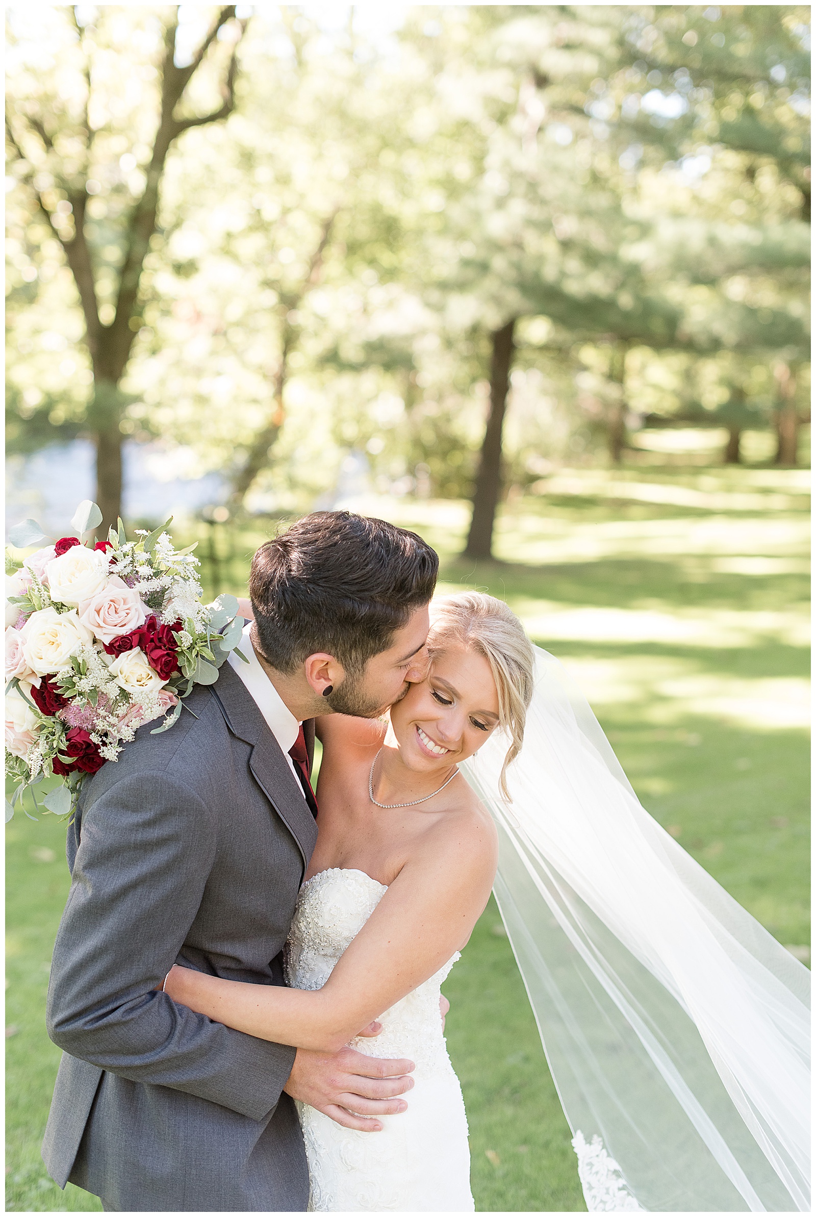 groom and bride are hugging with the groom on the left and the bride on the right and he is kissing her on the right side of her face while she holds her bridal bouquet around his back and looks down towards the ground smiling and her veil blows in the wind beside her and the groom is in a dark gray suit and the bride is in a white strapless gown with grass and trees and sunshine behind them at Riverdale Manor in Lancaster, PA