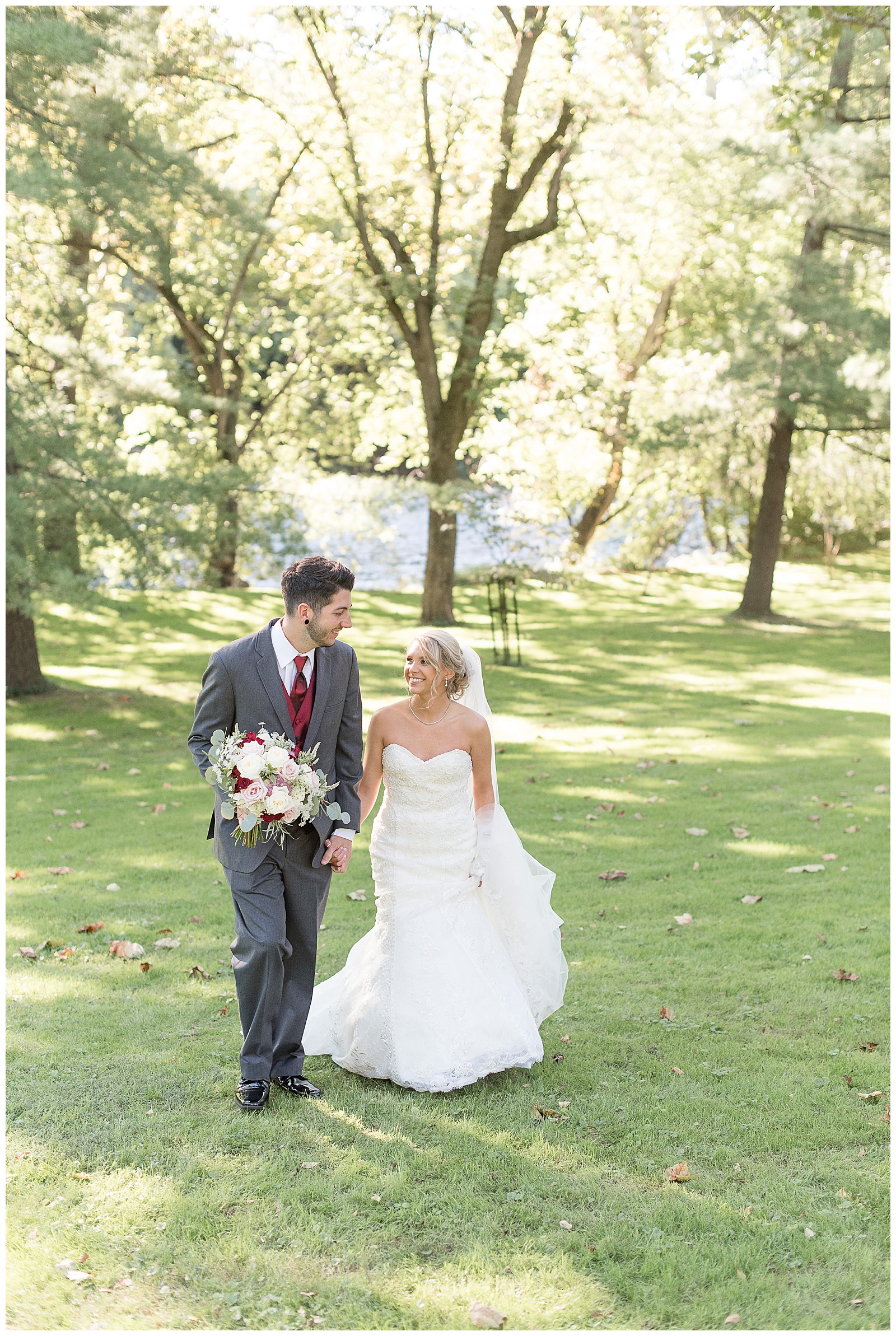 couple is holding hands and walking towards the camera smiling with groom on the left and bride on the right holding her gown with her left hand and the groom is holding the bride's floral bouquet in his right hand as they look at each other with grass, trees, and sunshine behind them at Riverdale Manor in Lancaster, PA