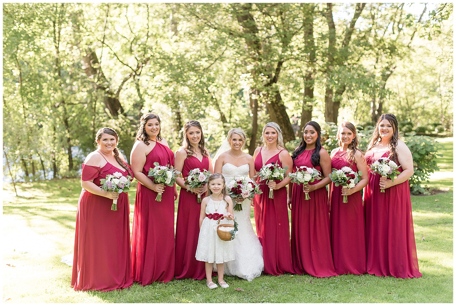 bride is standing in the middle of photo with three bridesmaids to her right and four bridesmaids to her left and the flowergirl standing in front of the bride and everyone is holding their floral bouquets and looking at the camera and smiling while standing in the grass with trees behind them at Riverdale Manor in Lancaster, PA