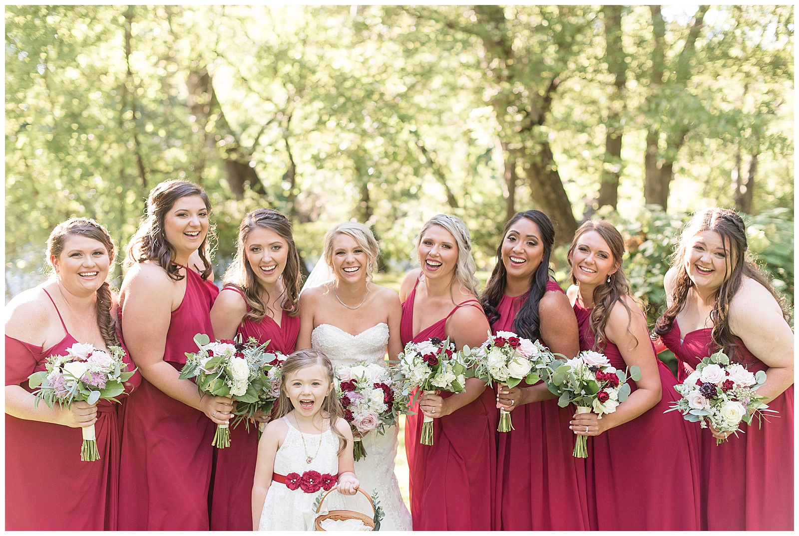close up photo of the bride with her bridesmaids and flowergirl all holding their flowers and smiling at the camera with trees and sunshine in the background at Riverdale Manor in Lancaster, PA