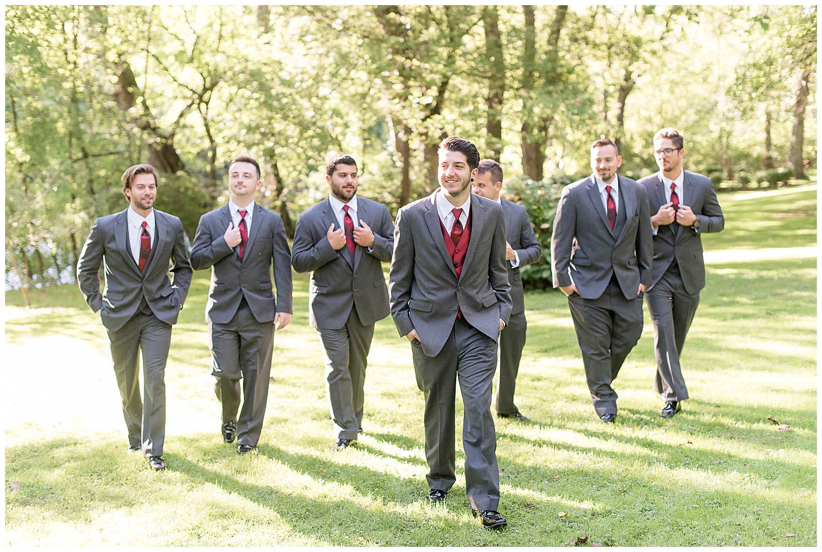 groom is standing in the center of the photo with his groomsmen slightly behind him side by side and they are all walking towards the camera with their hands either in their pockets or holding the edges of the suit coat near their tie and everyone is looking in different directions while smiling with trees and sunshine behind them at Riverdale Manor in Lancaster, PA