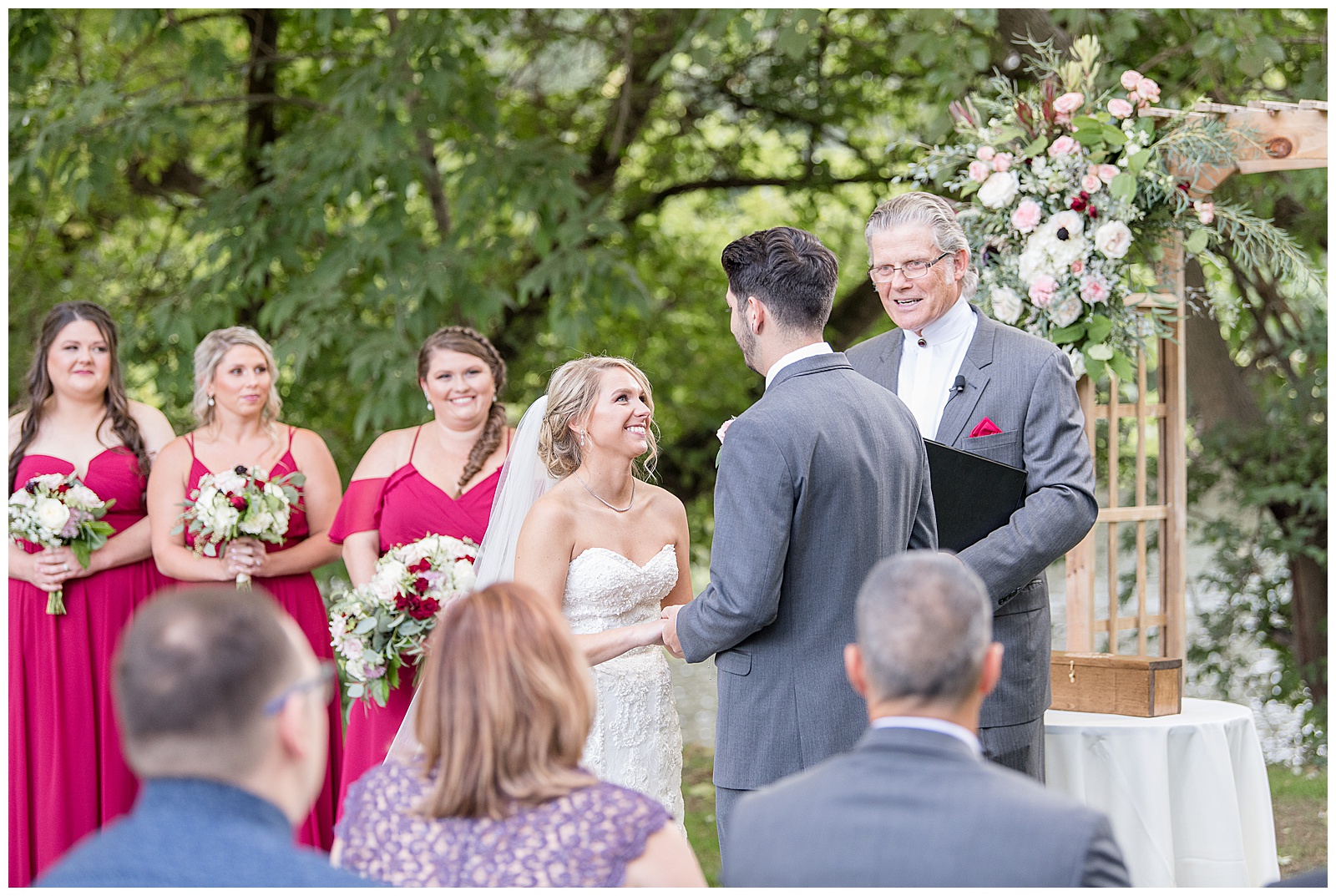 couple is facing each other with the bride on the left and her bridesmaids behind her the groom on the right and the officiant behind them and the couple is smiling and everyone is looking at them and smiling and there are guest seating at the bottom of the photo with their backs to the camera and trees are in the background and wooden archway with flowers and table with white linen tablecloth with trees in the background at Riverdale Manor in Lancaster, PA