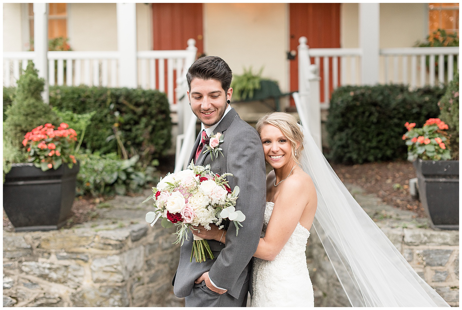 couple is standing in front of white house with two red front doors and the are facing the left side of the photo with the bride hugging the groom from behind and he is on the left and she on the right with her arms wrapped around him with her flower bouquet and she is looking at the camera smiling and he is looking back over his left shoulder smiling at Riverdale Manor in Lancaster, PA
