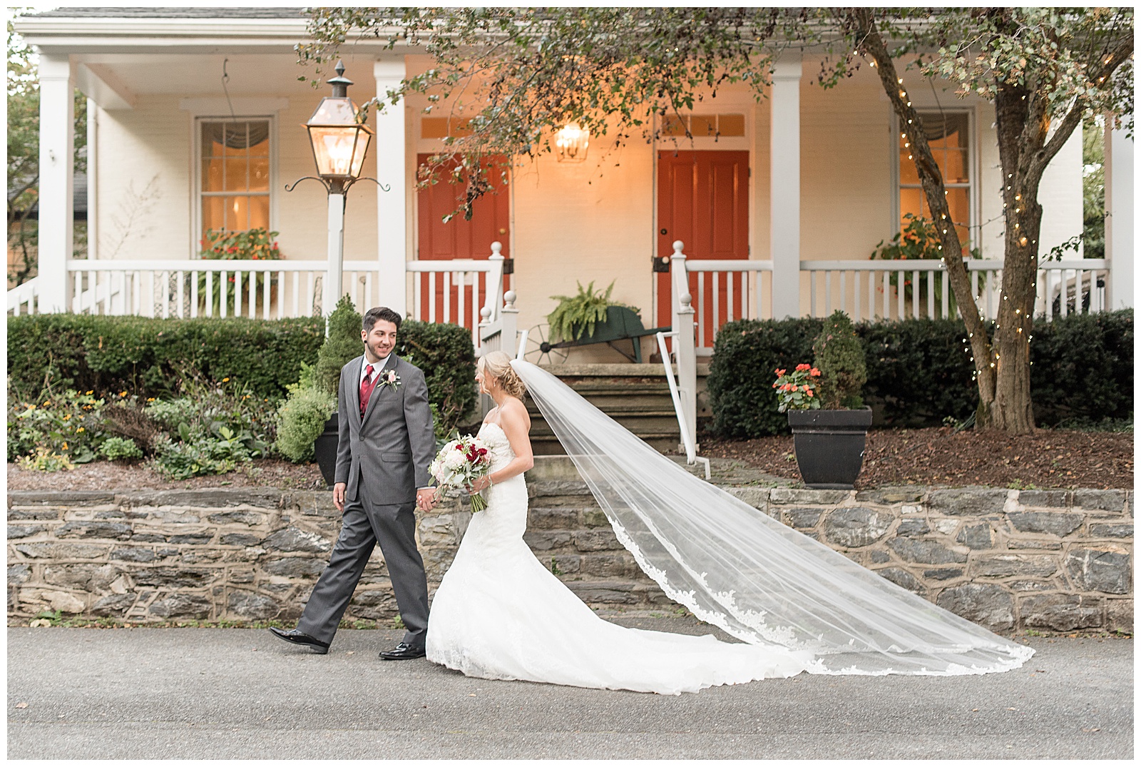 couple is walking on paved driveway in front of white house and the groom is to the left holding the brides right hand and leading her while looking back over his shoulder at her and smiling and the bride is on the right following behind him with her dress and veil extended behind her on the driveway and she is holding her flower bouquet at Riverdale Manor in Lancaster, PA