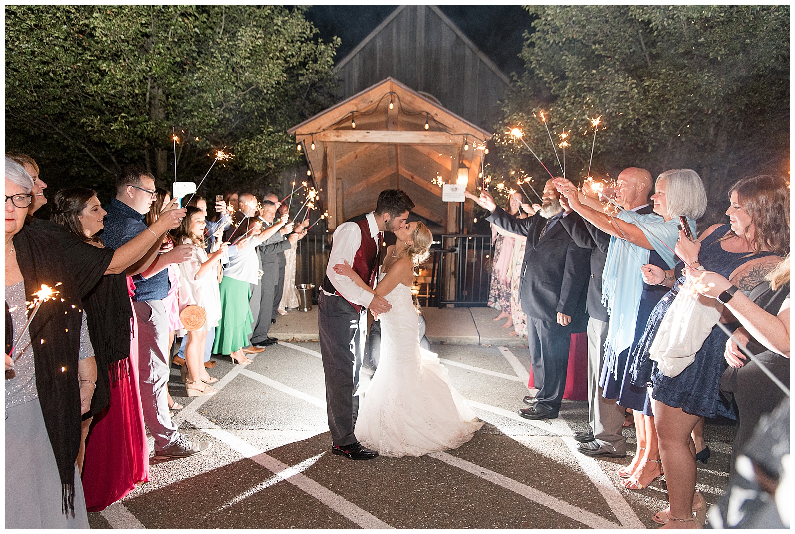 the bride and groom are kissing as they are leaving their wedding and reception and they are standing in between two long rows of guests who are extending sparklers overhead as the groom is on the left kissing his bride with a beautifully lit building and trees behind them at nighttime at Riverdale Manor in Lancaster, PA