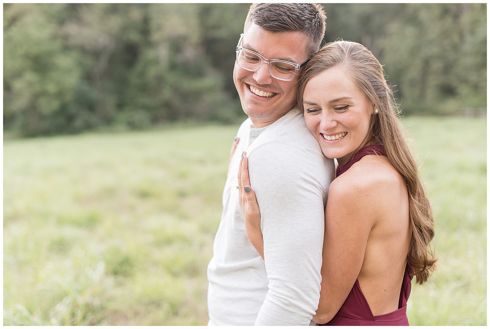 close up photo of the couple standing in grass field with trees behind them and they are both facing the left side of the photo with the guy on the left and his back to the girl and she is resting her right side of her face against the guy's back and left side of his face and they are both smiling and looking down towards the ground and the girl has her arms wrapped through the guy's arms at Hibernia County Park in Chester County, PA