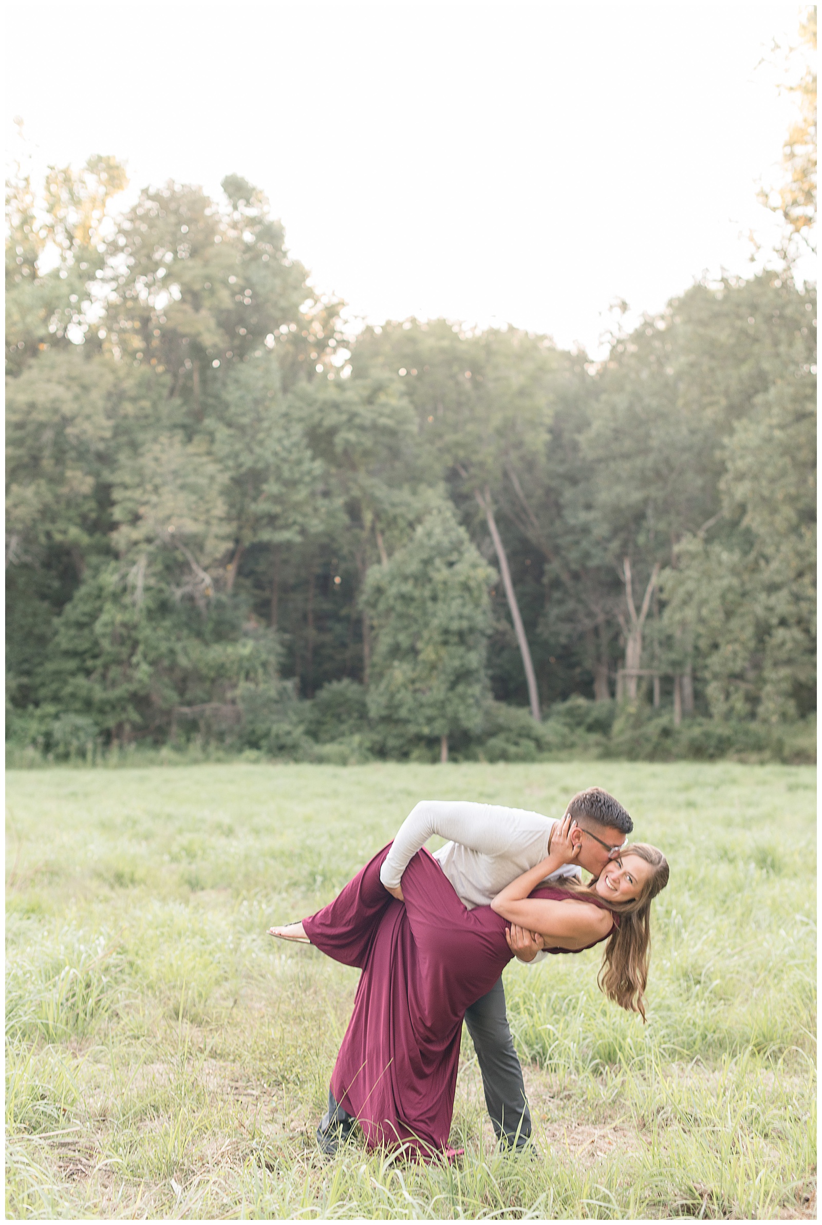 couple is standing in grass field with trees behind them and the guy is dipping the girl backwards and kissing her on her right cheek and holding her left leg up as the girl is wrapping her arms around the guy and looking at the camera and smiling at Hibernia County Park in Chester County, PA