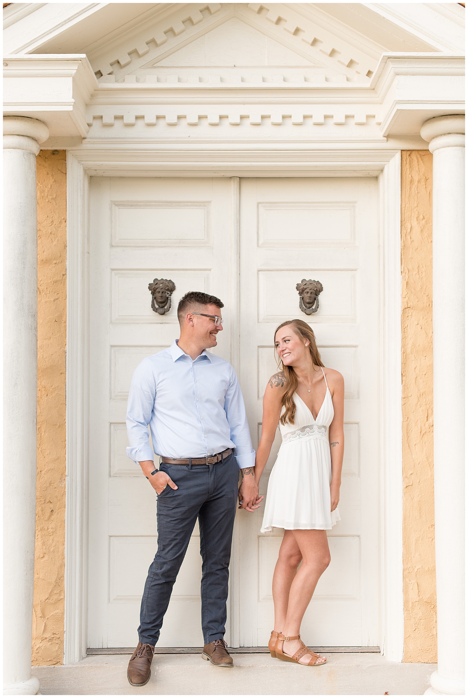 couple is standing in front of white doors of peach colored building and the guy is on the left with his right hand in his pocket and the girl is on the right with her left hand by her side and they are holding hands in the middle while looking at each other and smiling at Hibernia County Park in Chester County, PA