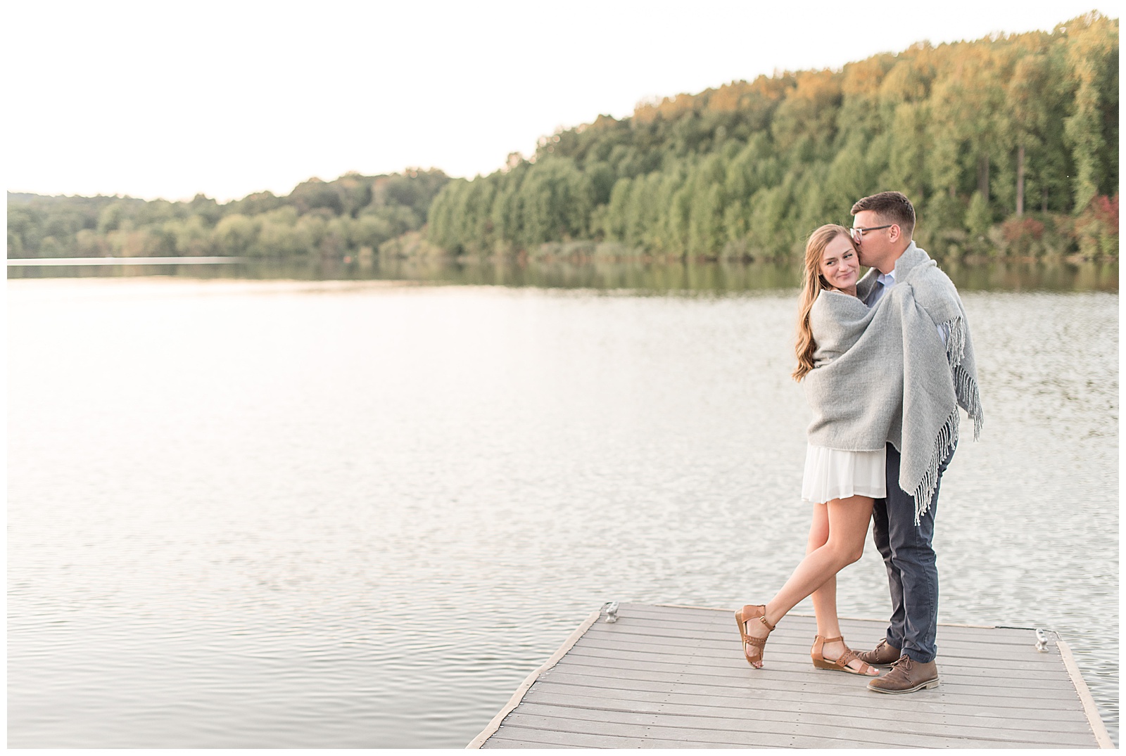 couple is standing at the end of dock with water and trees and sky behind them and the girl is on the left and the guy is on the right and they are facing each other hugging tightly with a gray blanket wrapped around them and she has her right leg propped back while looking towards the camera as he is kissing the left side of her head at Hibernia County Park in Chester County, PA
