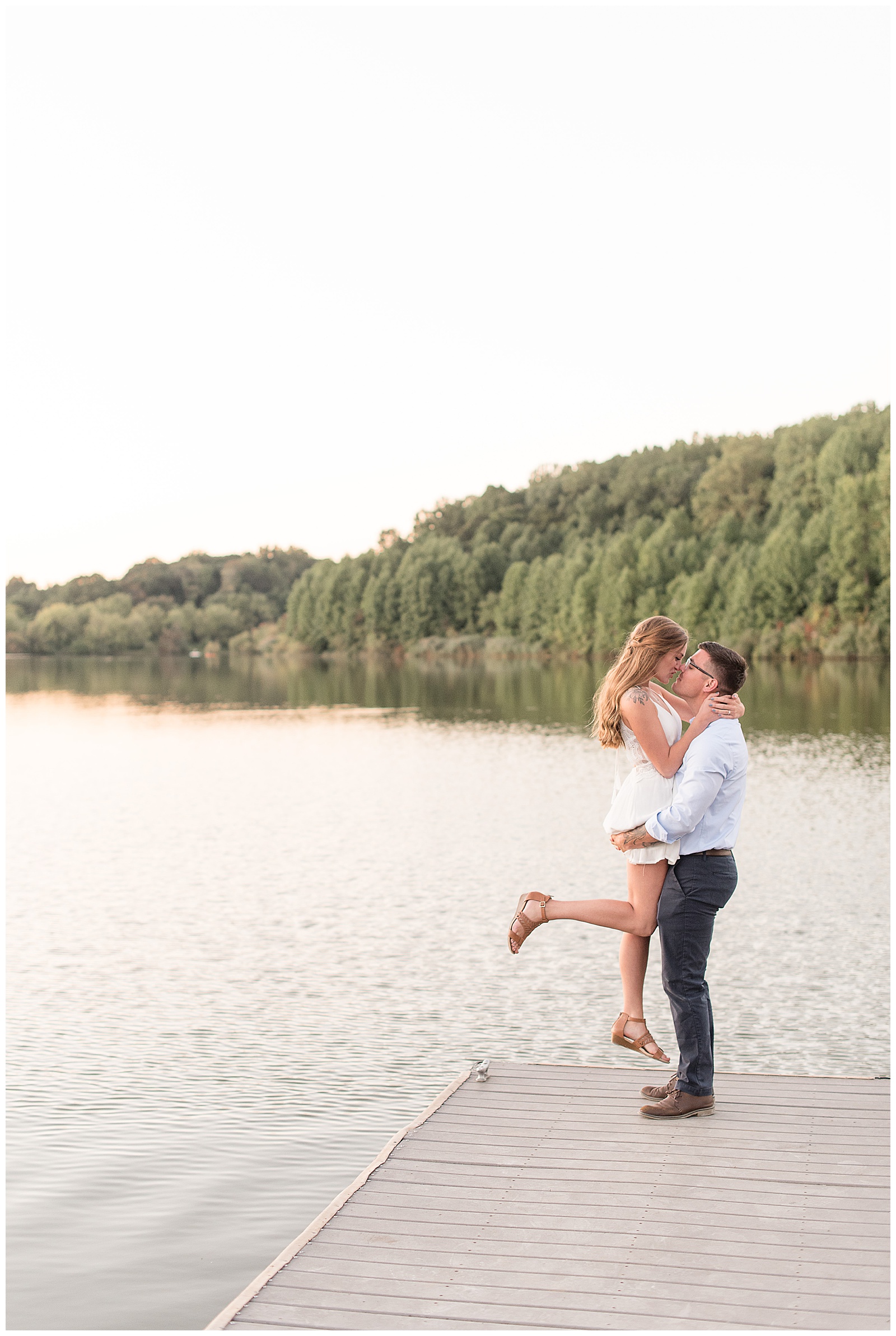 couple is standing at the end of dock with water and trees and sky behind them and the guy is on the right and is lifting the girl up as she bends her right leg and they have their arms wrapped around each other at Hibernia County Park in Chester County, PA