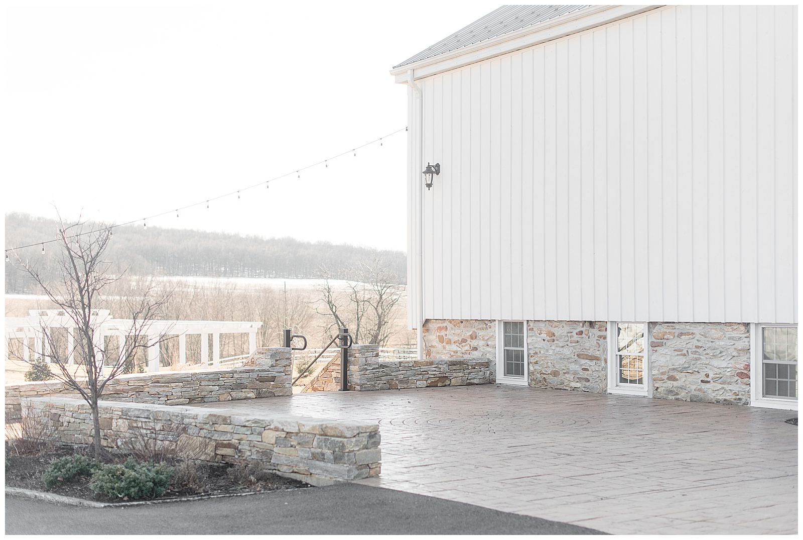exterior photo of corner of large white barn with fields and mountains and skyline off in the distance and nice stamped concrete patio with stone wall around it and white windows and stone exterior along bottom of barn at The Barn at Stoneybrooke in Atglen, PA