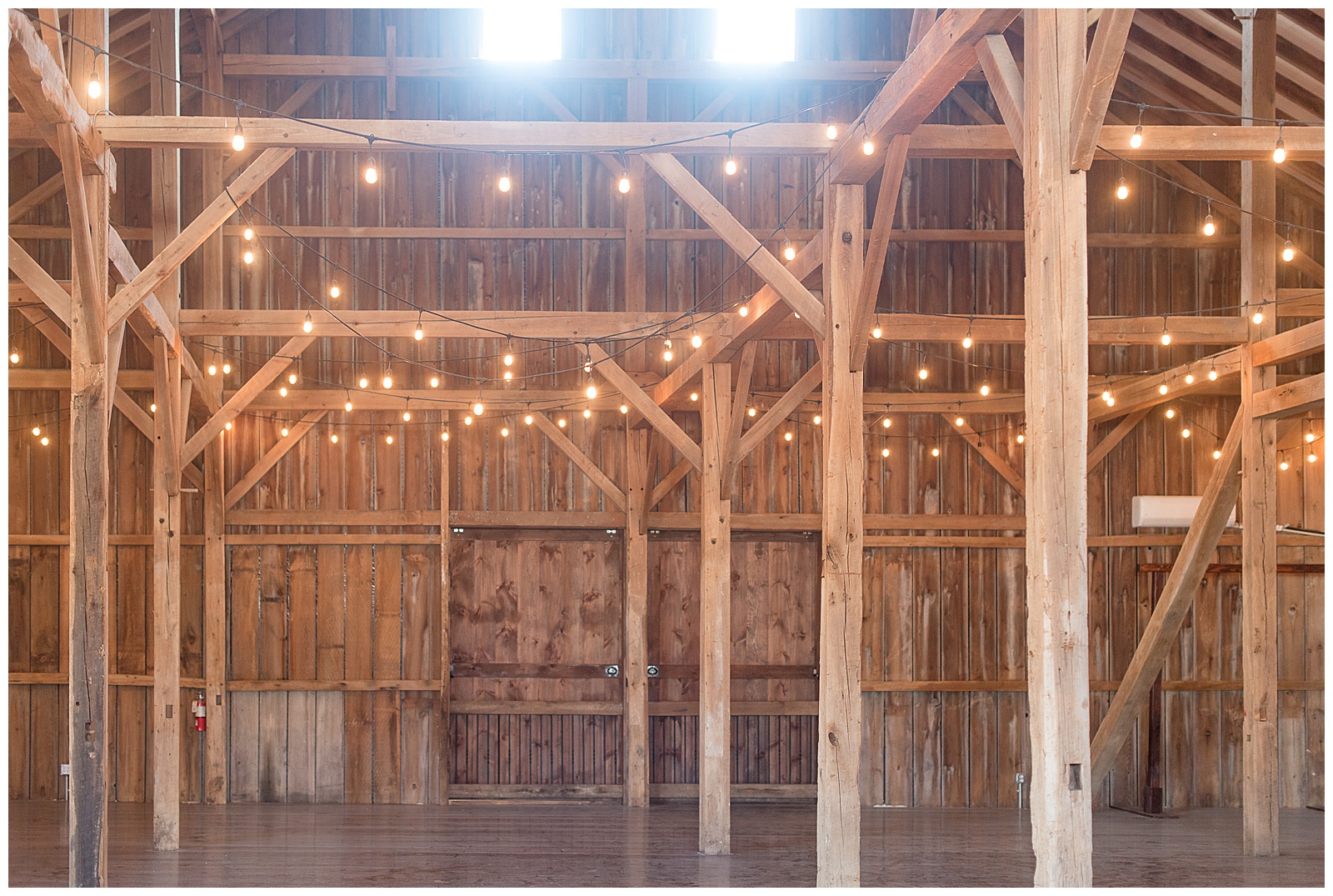 interior photo of exposed wooden beams and walls of barn reception area with several strings of lights hanging from rafters with the light of the outdoors shining in from two windows at the top of the photo at The Barn at Stoneybrooke in Atglen, PA