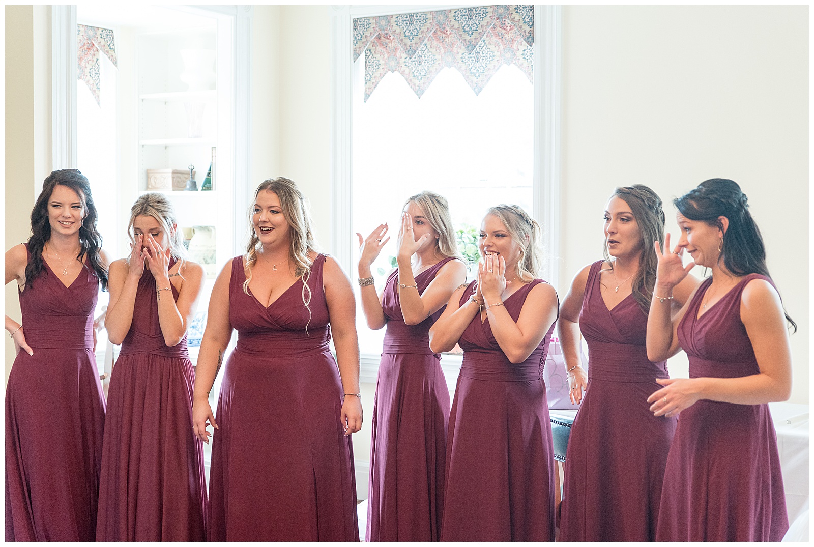 bridesmaids in maroon dresses reacting as they see bride for first time in bridal suite