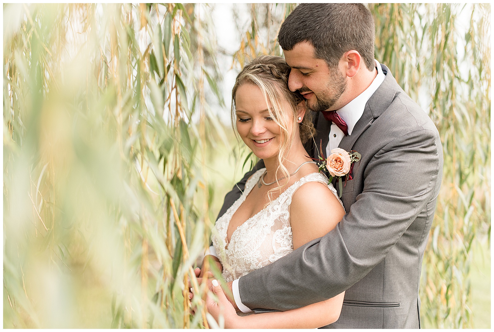 groom hugs bride while smiling under willow tree at hayfields country club in maryland