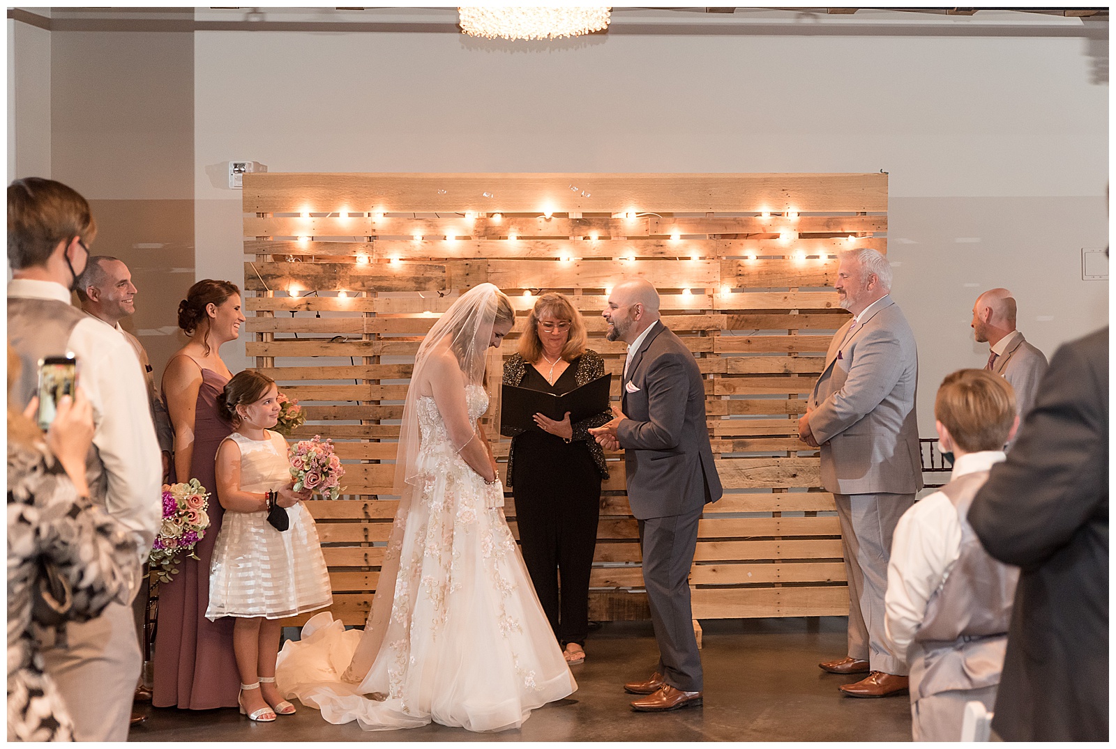 couple standing with bridal party and officiant during wedding ceremony by wooden wall with twinkling lights