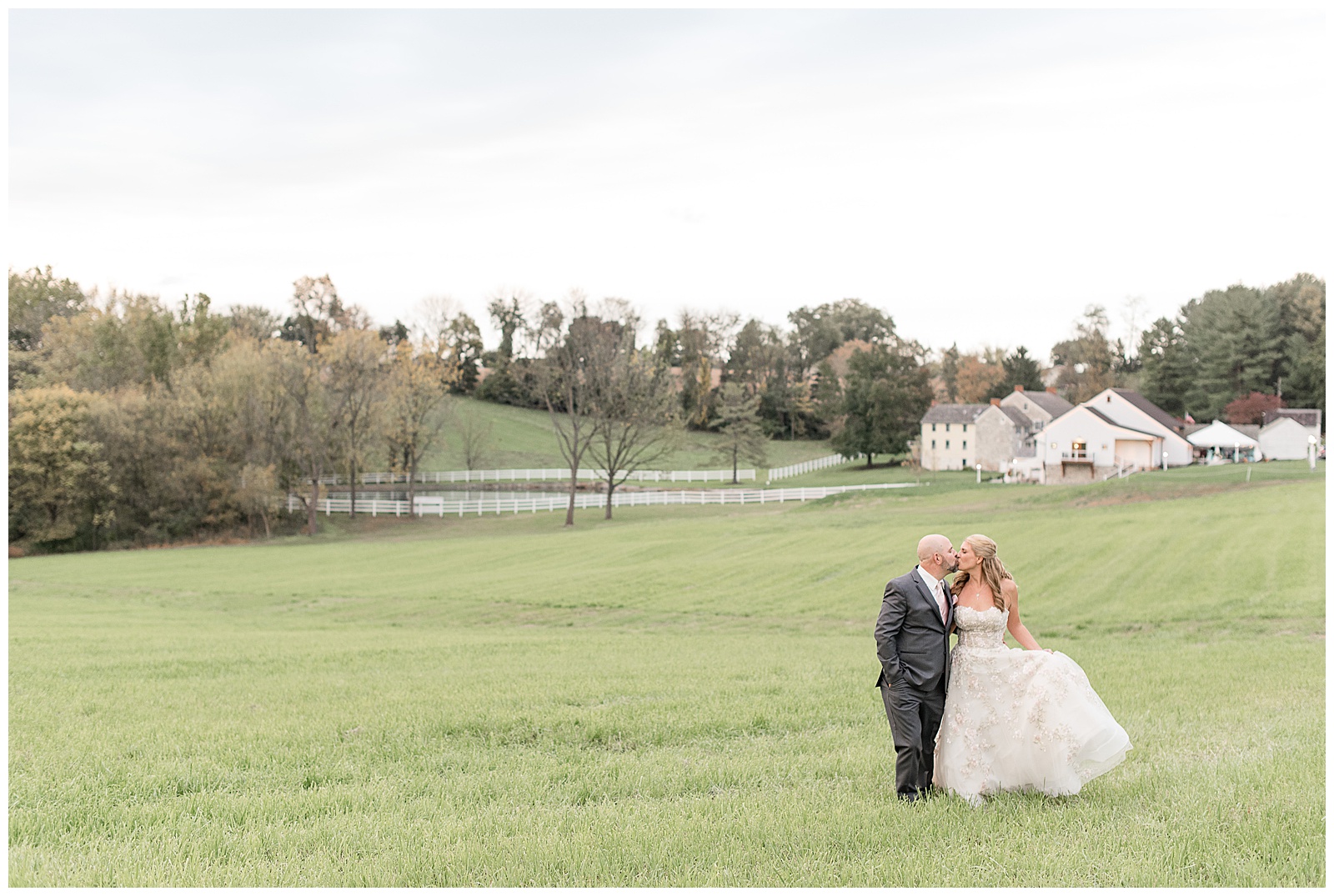 couple kissing in meadow with white fence and barn and trees in background at bluestone estate