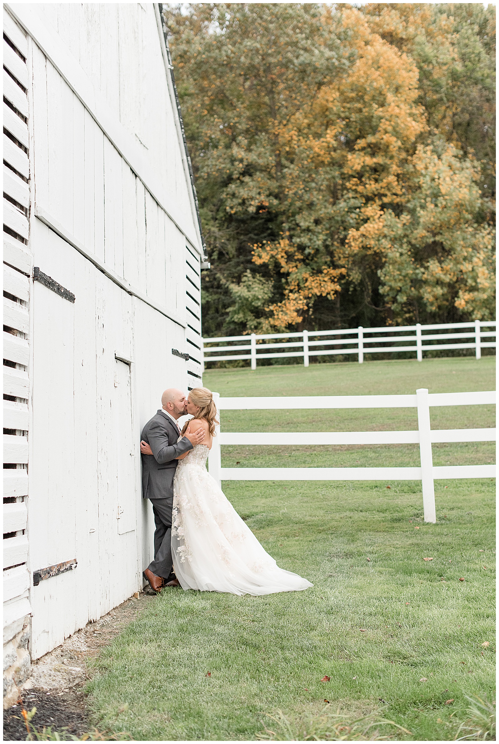 groom and bride leaning against white barn kissing by fence and fall trees in lancaster pennsylvania