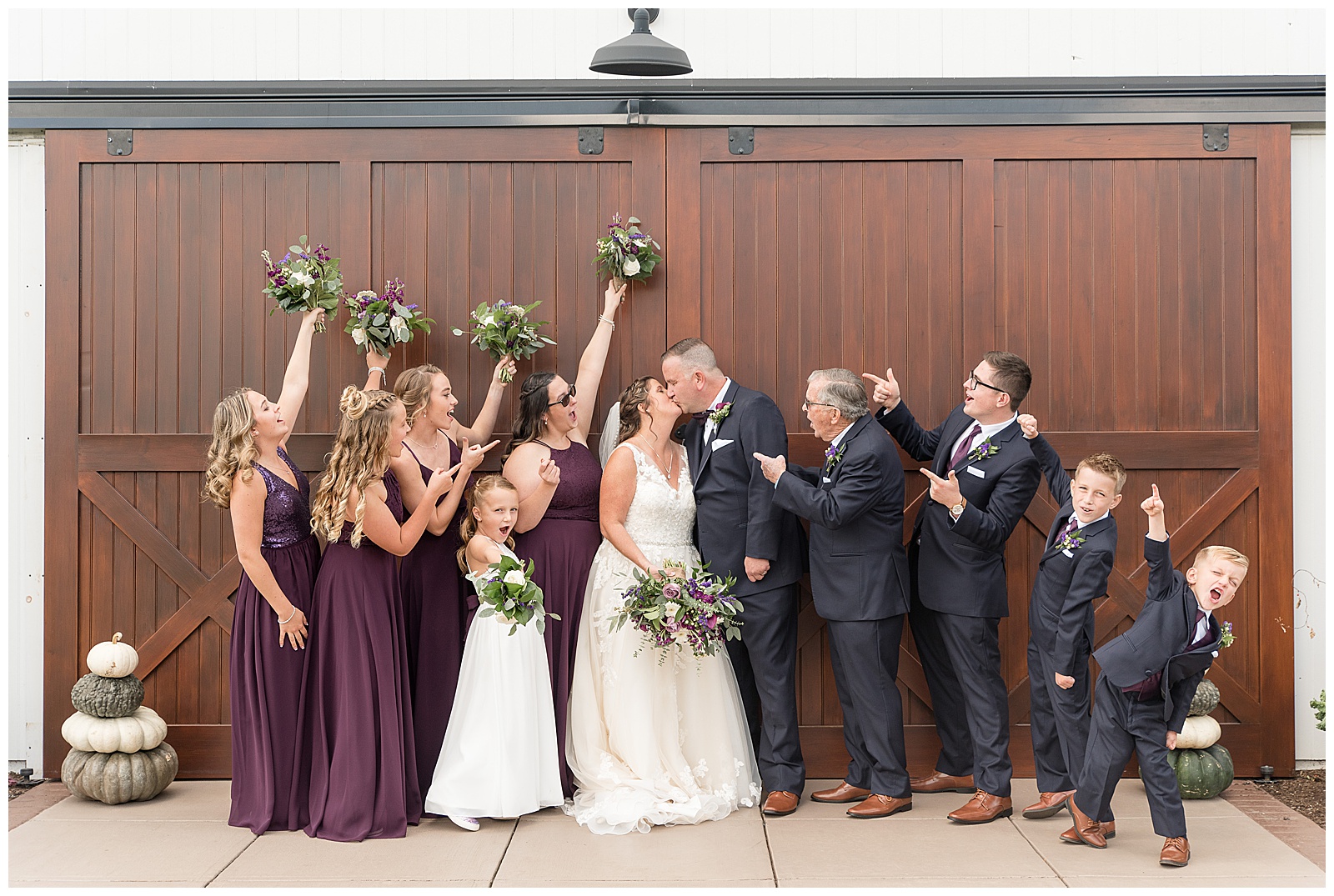 Full Bridal party cheering as bride and groom share a first kiss in front of large wood barn doors