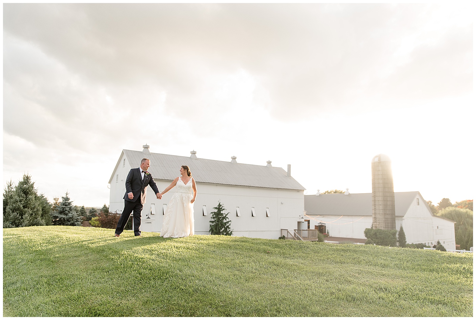 bride and groom walking on grass mound overlooking venue with sun setting behind them