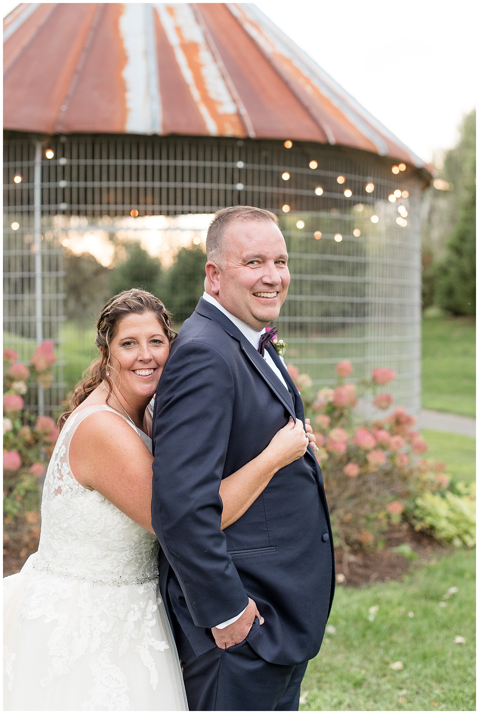 wife has arms wrapped around groom from behind as they both smile towards camera with corn crib behind