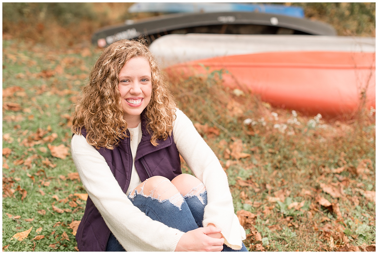 senior girl sits on grass hugging her knees by colorful canoes at speedwell forge lake