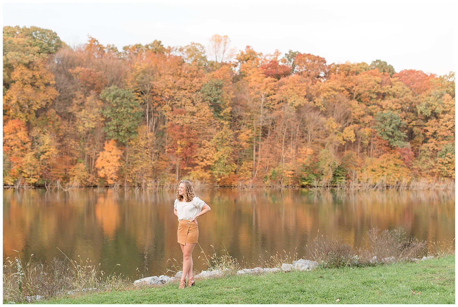 senior girl hands on hips by colorful trees at lake in lititz pennsylvania