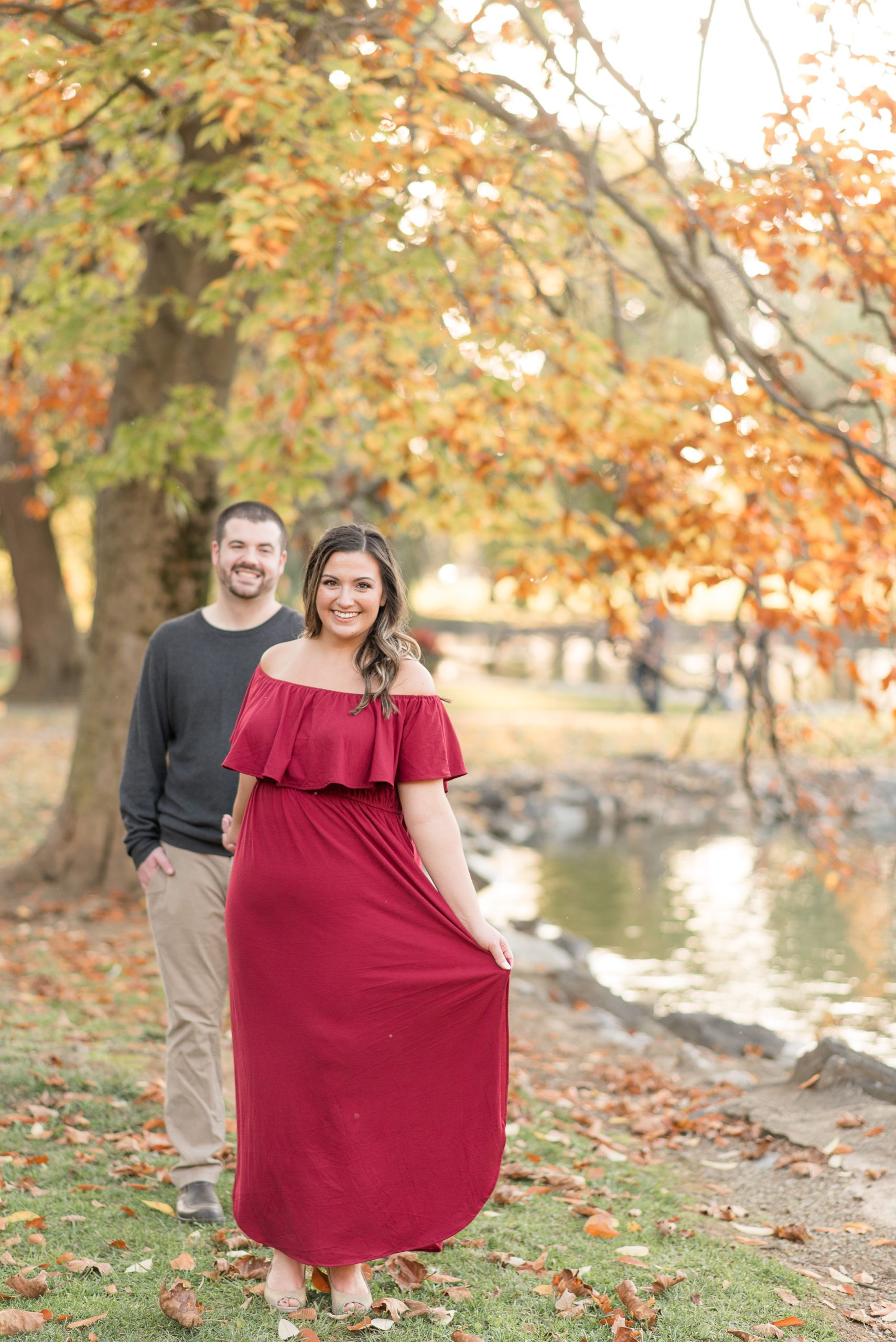 couple smiles as girl extends her dress by pond with beautiful fall leaves at long's park