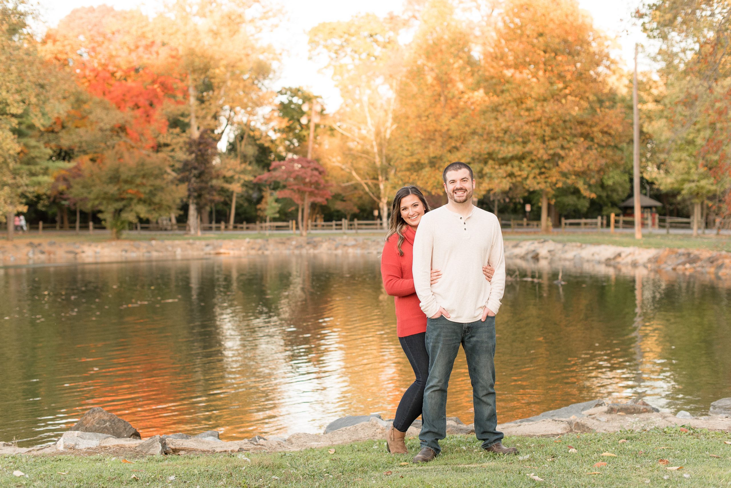 girl hugs guy while standing by pond with beautiful fall trees behind them at long's park