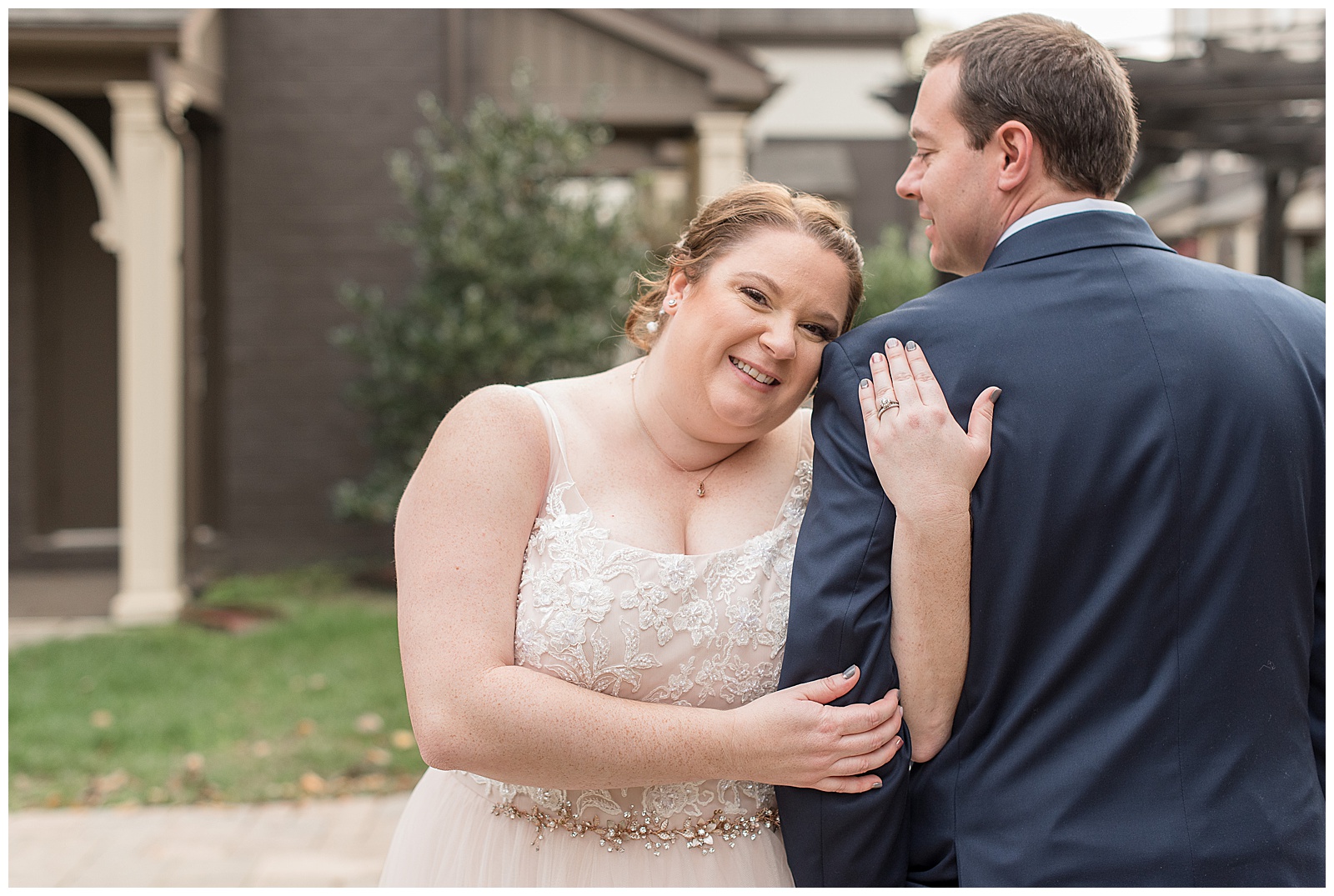 bride smiling wrapping her arms around groom's left arm with his back to camera outdoors