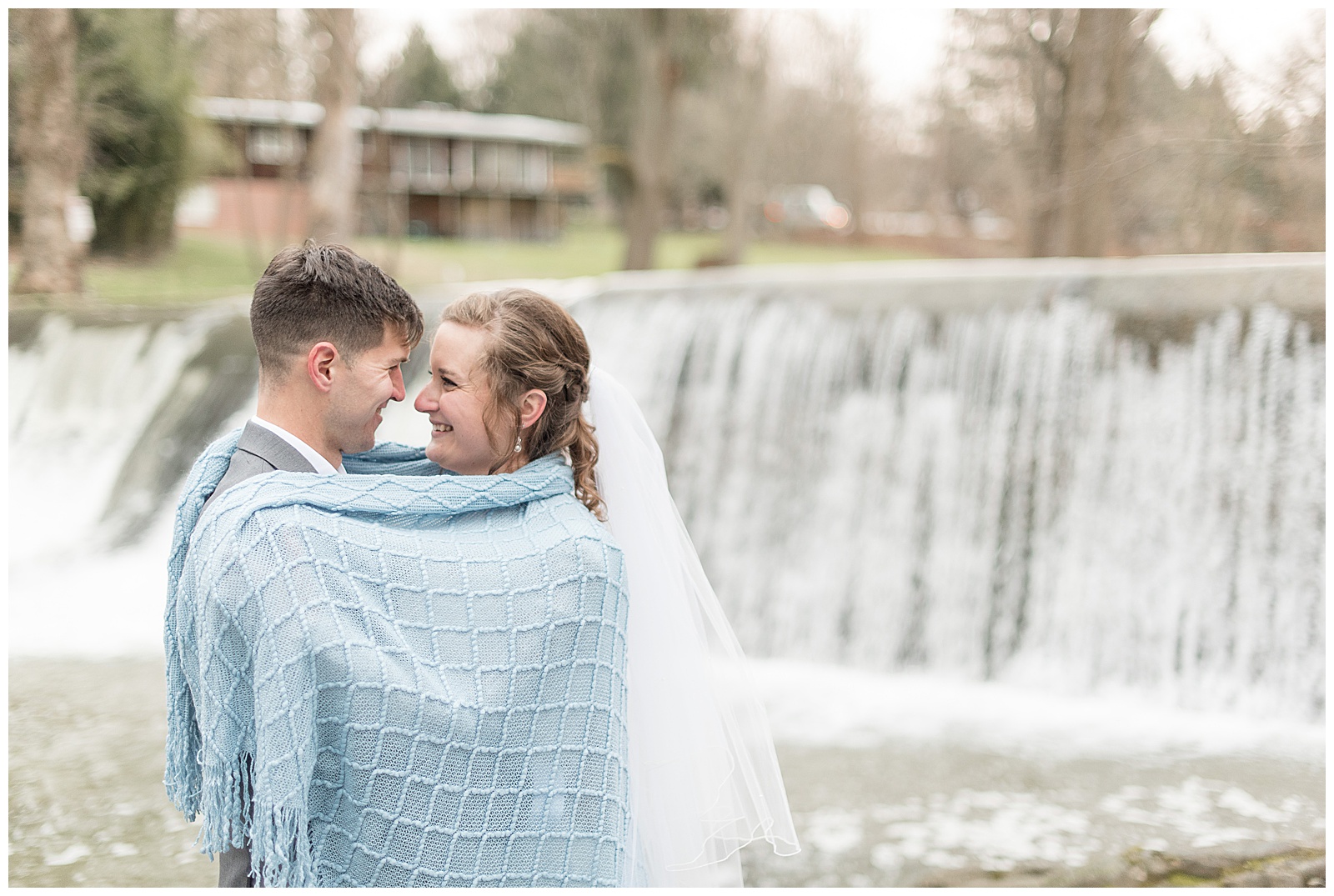 bride and groom snuggle under light blue blanket by waterfall at the mill at manor falls