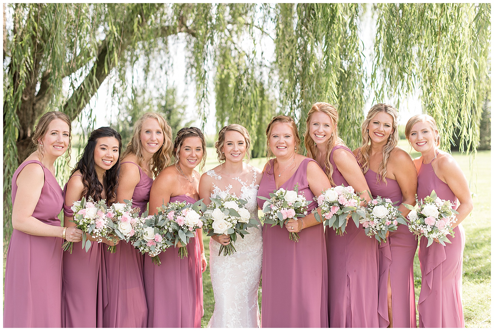 bride standing with her eight bridesmaids in mauve dresses all holding bouquets under willow tree
