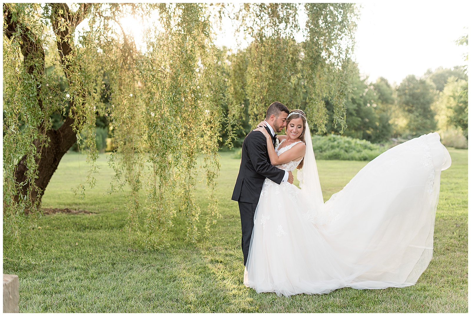 bride and groom hug as bridal gown train extends into the air behind her as they stand under willow tree