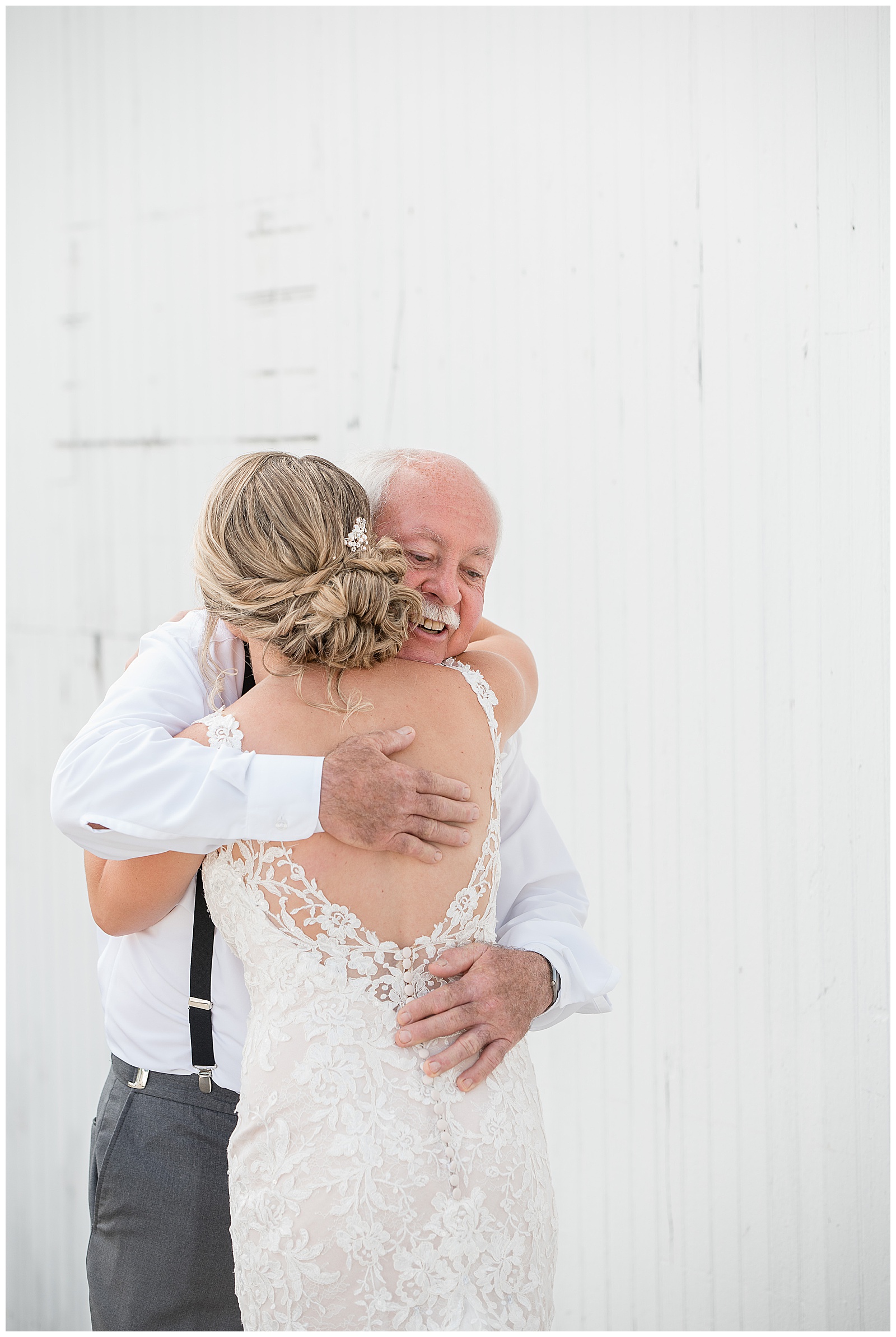 father of the bride smiling and hugging the bride before her wedding day along white barn wall