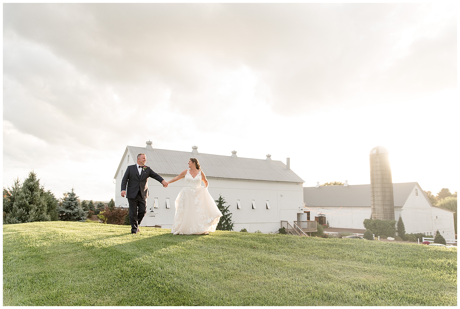 bride and groom holding hands walking down grassy hill with white barn in background at stoltzfus homestead and gardens