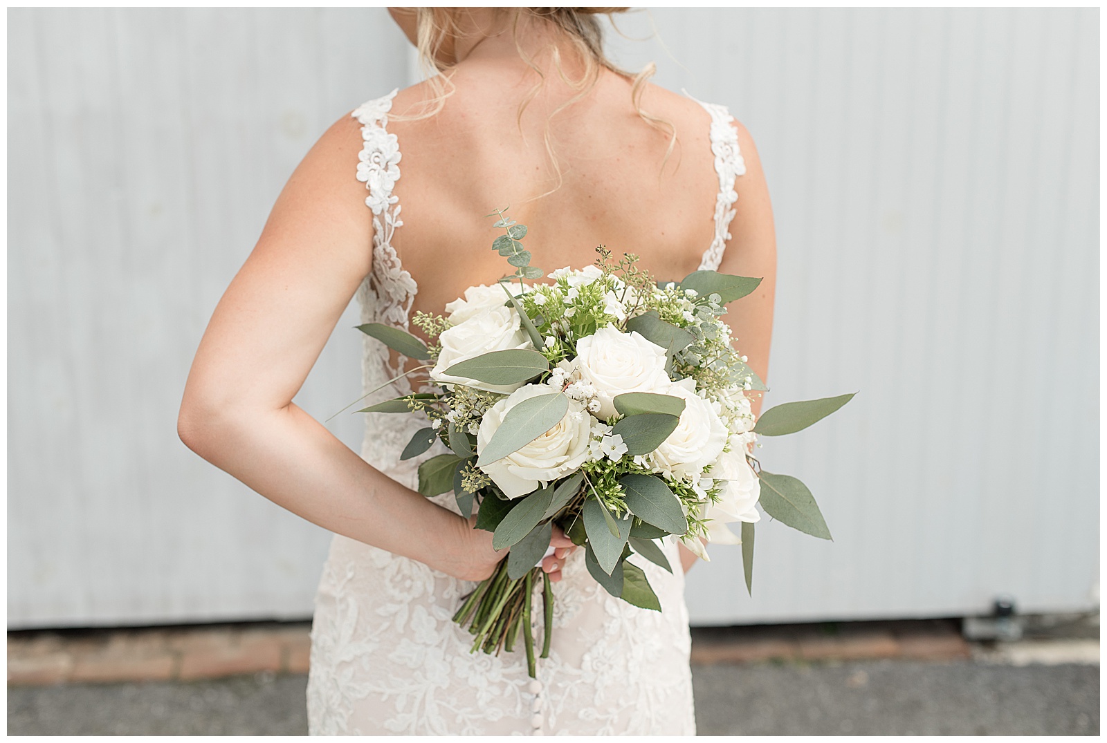 bride's back to camera holding bouquet behind her back with variety of white flowers and eucalyptus by white barn door