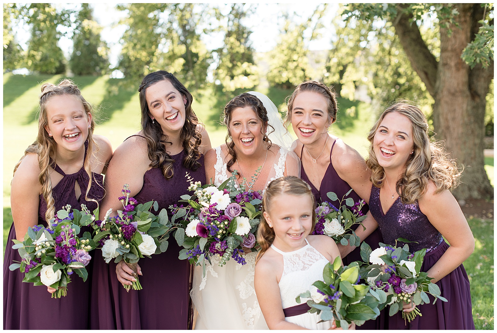 bride surrounded by bridesmaids and flower girl all huddling together with bouquets and leaning in smiling joyfully