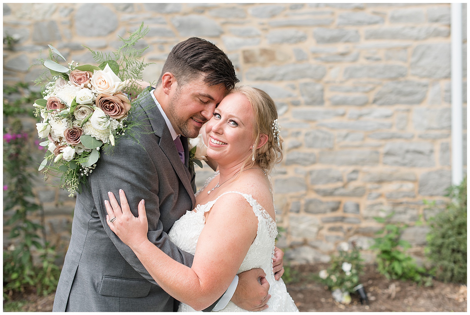 groom hugs bride tightly as she smiles at camera by stone wall at fall tree farm bed and breakfast