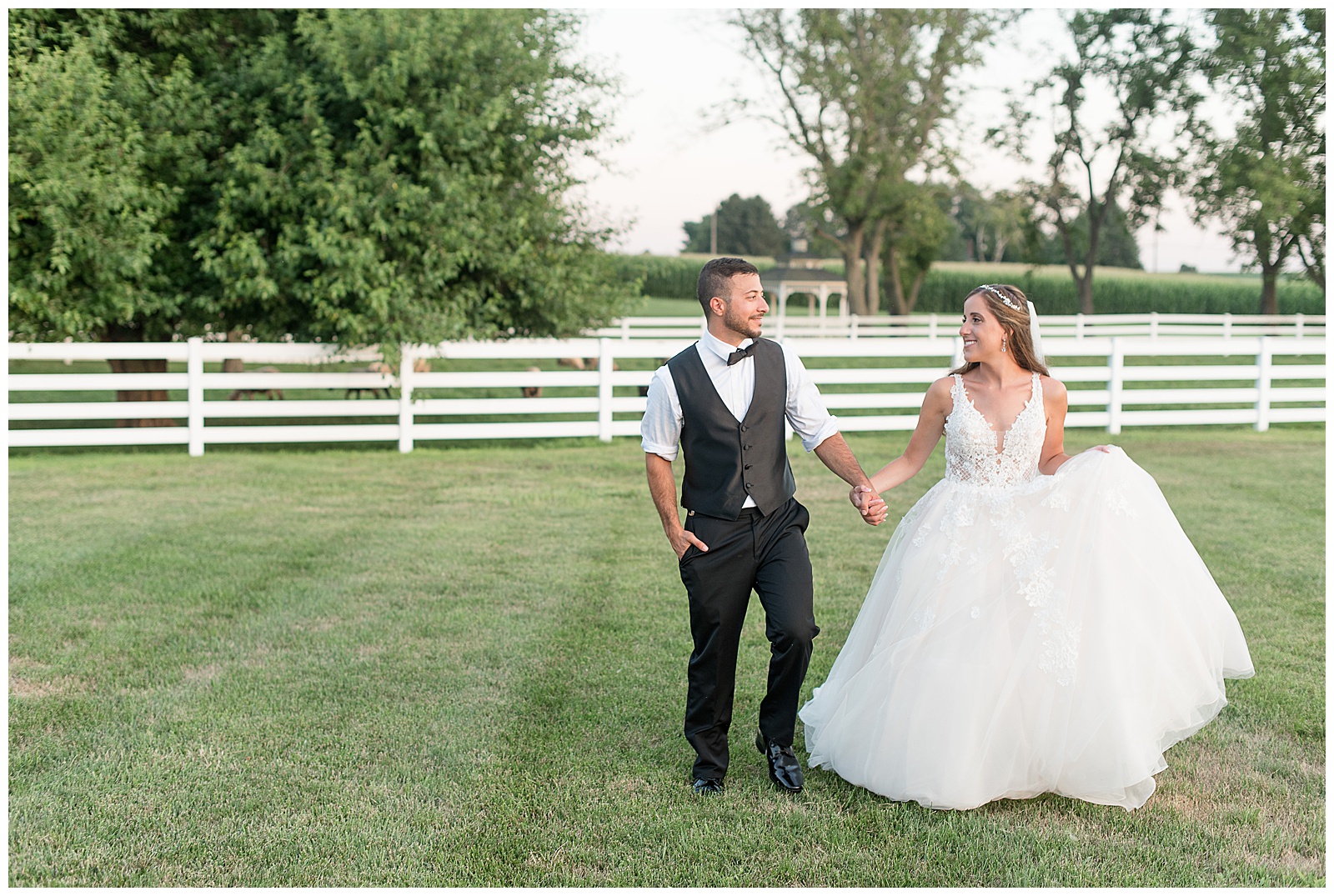bride holding grooms hand leading him as they smile and look at one another in grass with white fence and trees behind them