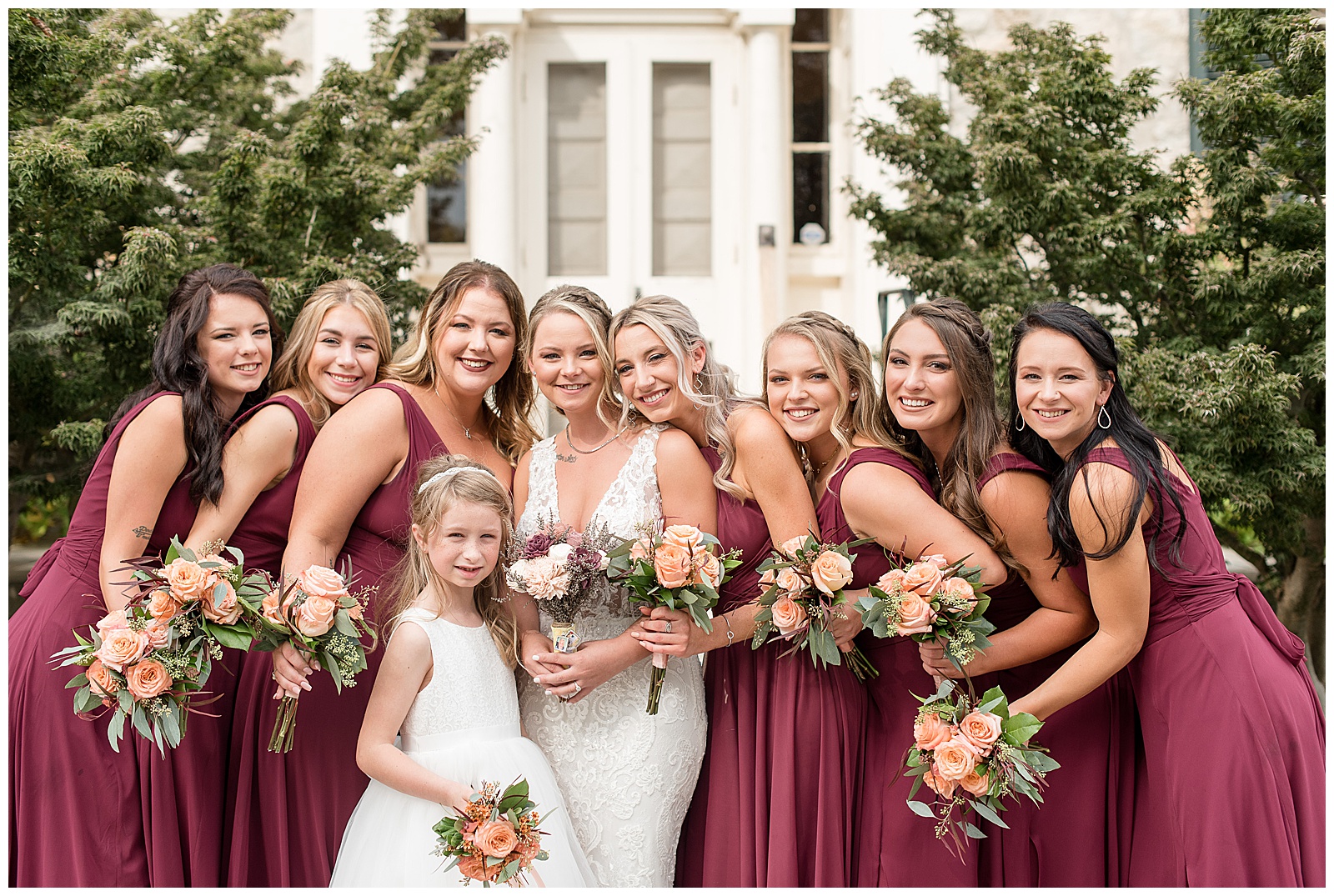 bride surrounded by her bridesmaids in maroon gowns and flower girl all holding bouquets outside white building