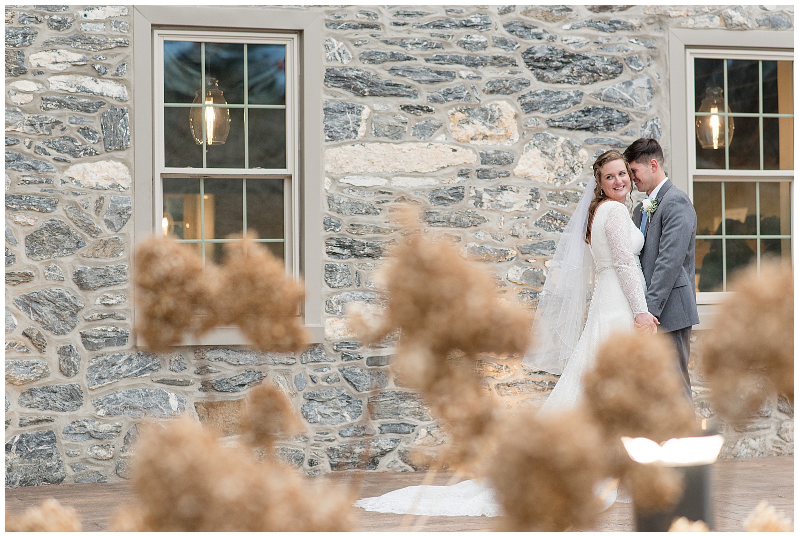 bride looking over shoulder smiling as groom smiles resting his forehead against hers by stone building with dried out hydrangeas in front of them